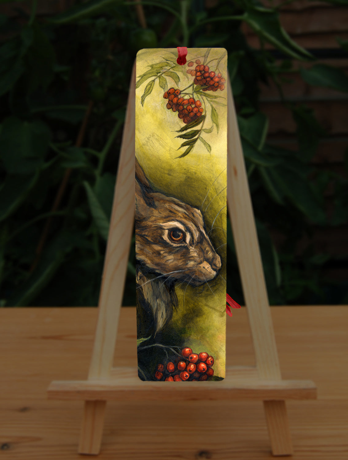 A rectangular bookmark: The head of a hare facing to the right. Sprigs of red rowan berries and leaves are drawn above and below. The background is mottled yellows and browns.