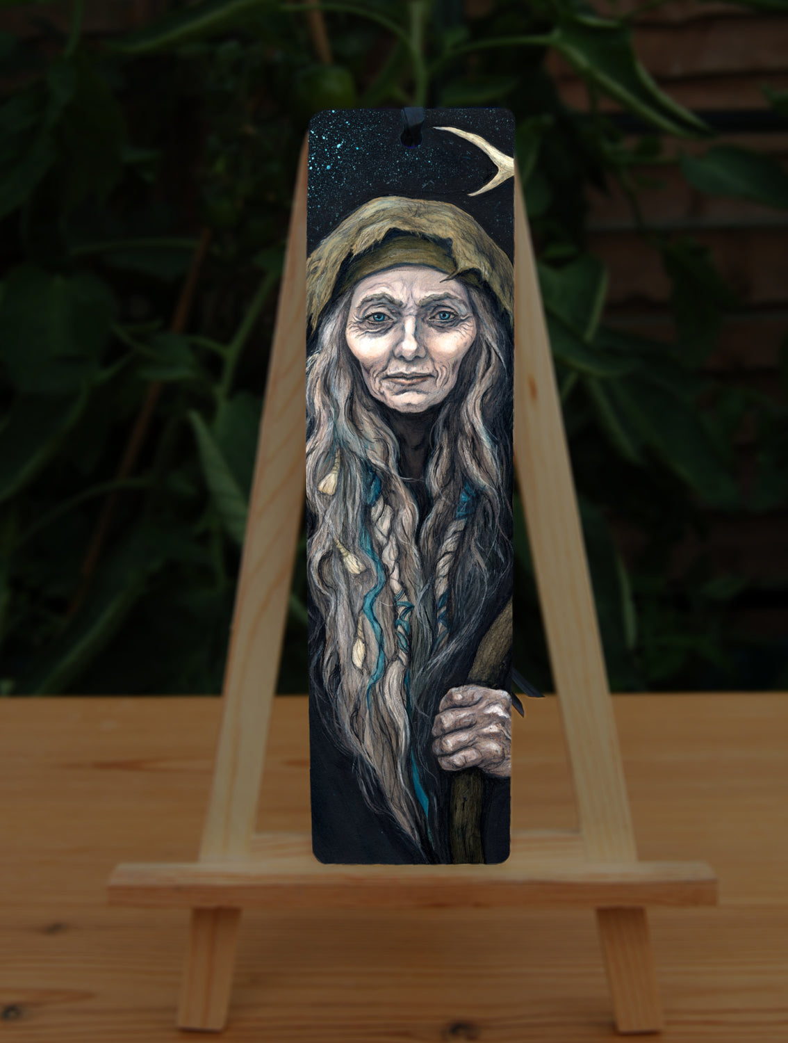 A tall bookmark: An older woman with blue eyes stands facing the viewer. Her head is covered by a brown hood, and her long grey hair, some of it braided with blue braid, falls down below her face. She holds a staff, topped with a deer antler, upright in one hand. A sea of stars shine behind her.