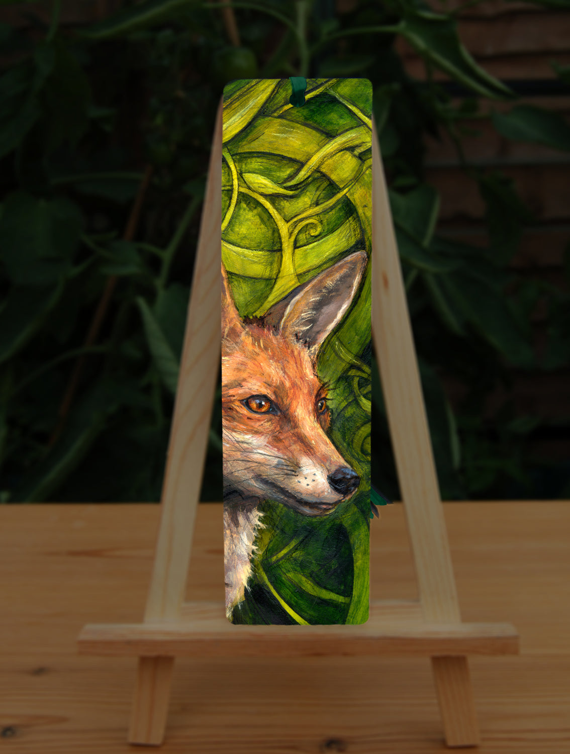 A tall bookmark: The head and neck of a red fox, their face angled towards the front right. The background consists of swirls of branches and leaves in a rich green colour.