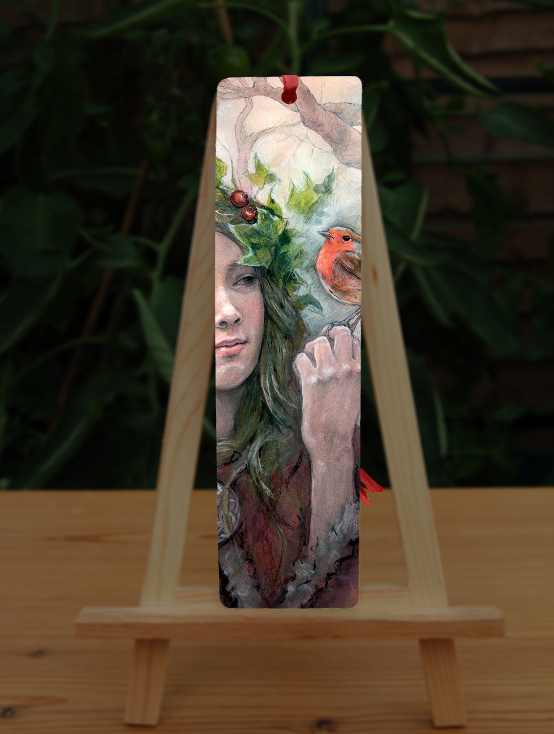 A rectangular bookmark: A young woman looks towards a robin sitting at eye level on the fingers of her hand. She wears a red cloak with white fur trim, closed with a large Celtic brooch. Sprigs of red berries and ivy are woven into her hair. Bare branches of trees fill the background.