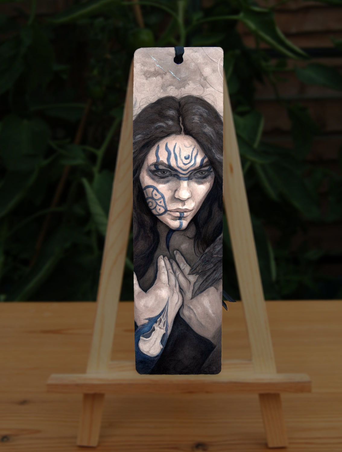A tall bookmark: The head and hands of a woman. She has long dark hair and her face is decorated with patterns in blue paint. Her hands are held together against her body. On her wrist she wears a blue band with a raven skull attached. The background is a mottled brown.
