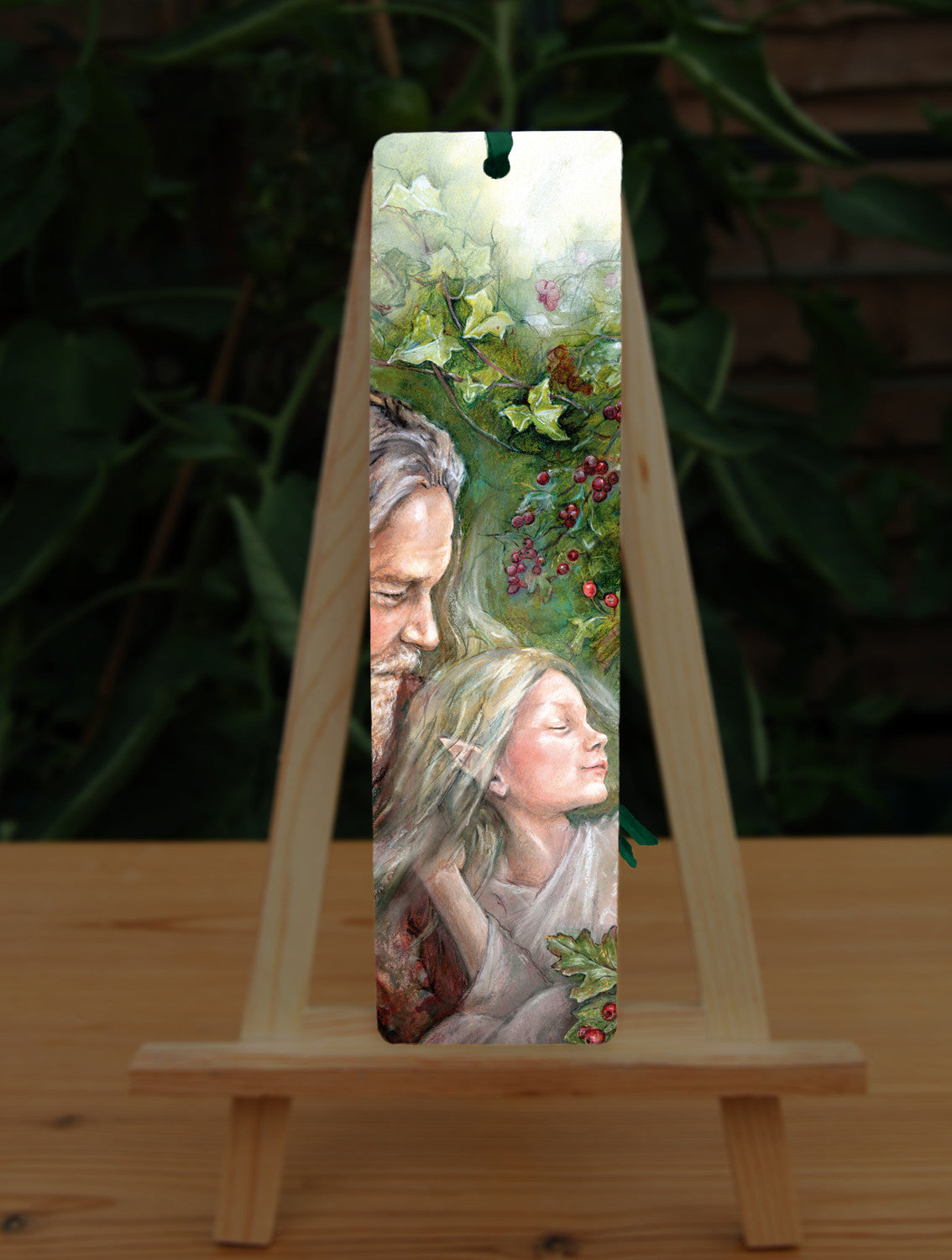 A rectangular bookmark: An older man with long hair, grey beard, and wearing a red coat cradles a sleeping fairy, wearing a pale gossamer dress, in his arms. Branches of hawthorn and ivy fill the background.