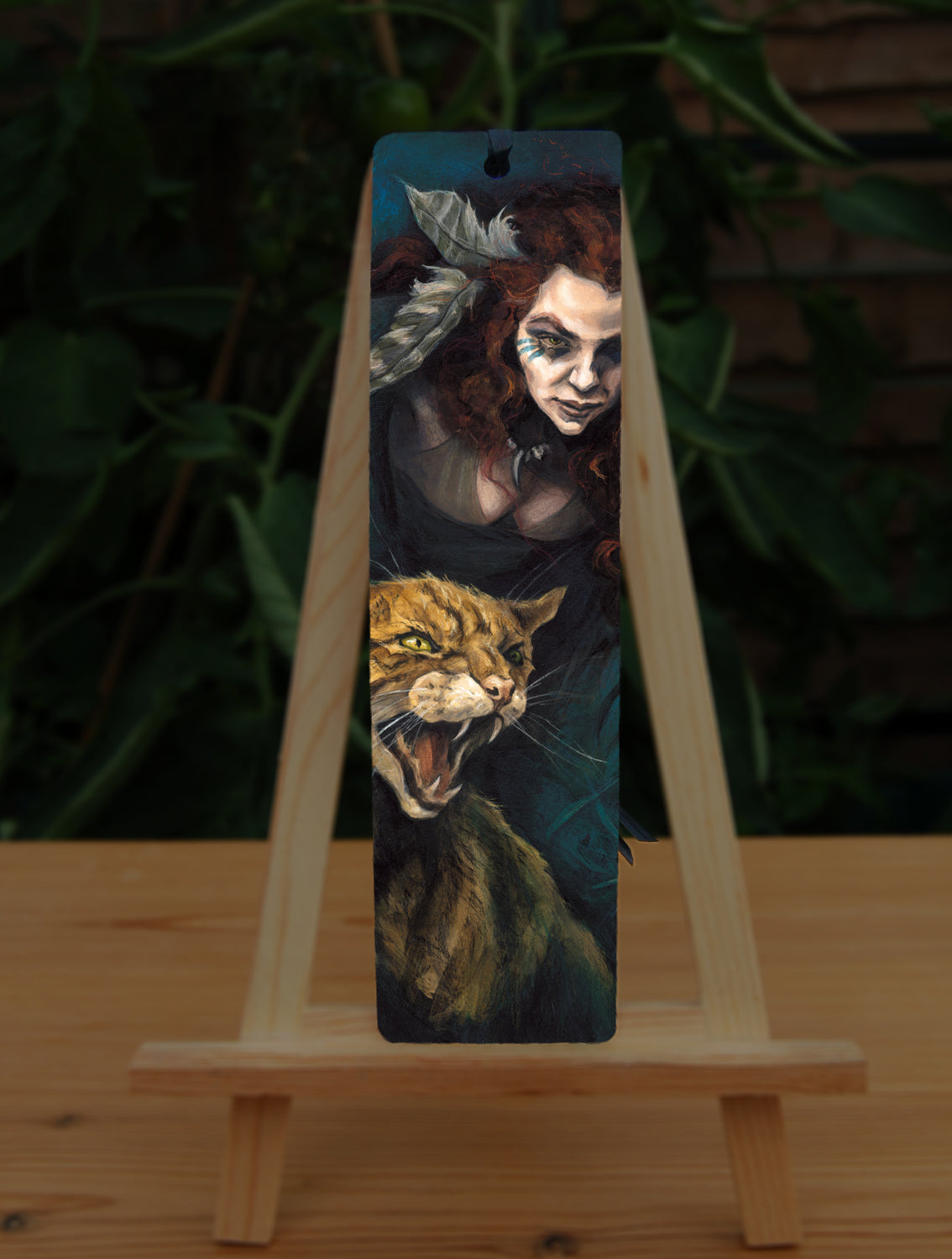 A tall bookmark: The head and chest of a woman with long red hair decorated with two feathers, and with lines of blue paint extending away from her eyes, looms into the picture above the head and body of a snarling brown striped cat. The background is teal and green vegetation.