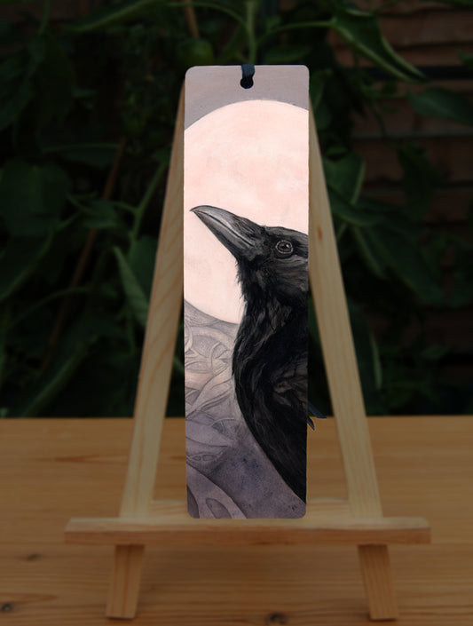A tall bookmark: The head and body of a raven standing facing towards the left, head tilted upward, behind which is a full moon. The background consists of swirls in a pale pink-brown colour.