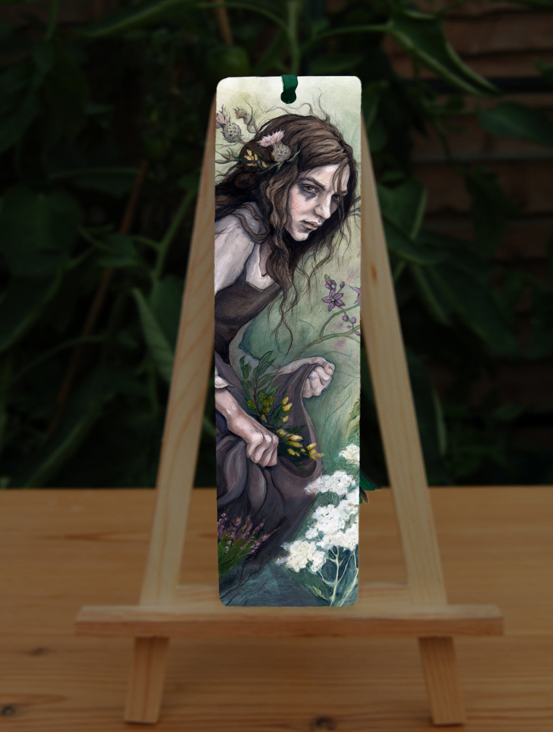 A tall bookmark: A young woman wearing a long brown sleeveless dress with a pale long sleeved top underneath, is surrounded by flowers as she kneels on the ground. The front of her dress is gathered in her hands to form an apron containing sprigs of bog myrtle and yellow gorse flowers. Heather grows next to her. Woody nightshade grows the other side with yarrow to the front. Thistles appear to be tied into her long brown hair.