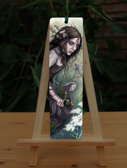 A tall bookmark: A young woman wearing a long brown sleeveless dress with a pale long sleeved top underneath, is surrounded by flowers as she kneels on the ground. The front of her dress is gathered in her hands to form an apron containing sprigs of bog myrtle and yellow gorse flowers. Heather grows next to her. Woody nightshade grows the other side with yarrow to the front. Thistles appear to be tied into her long brown hair.