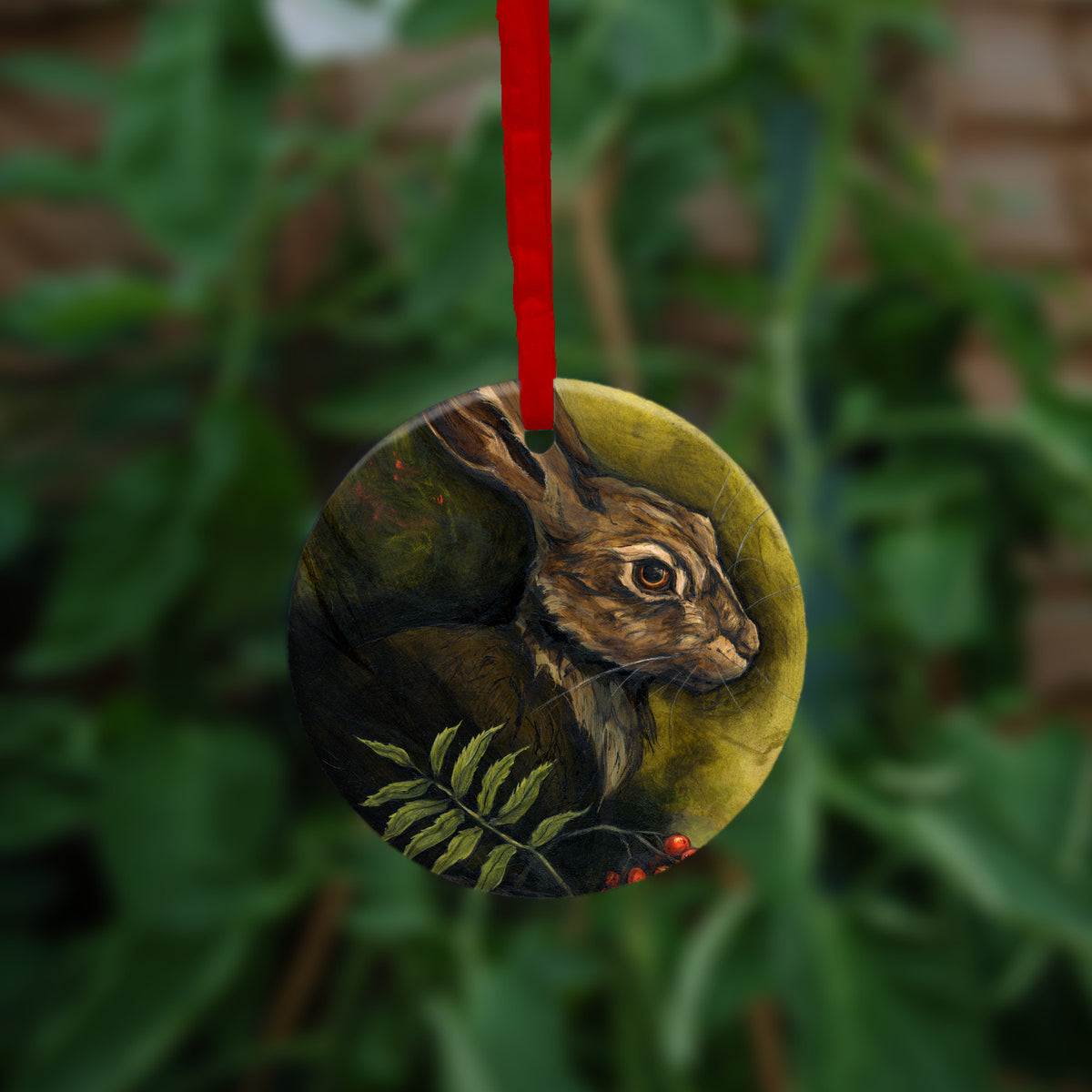 Circular hanging ornament with a red ribbon: The head and shoulders of a hare facing to the right. Sprigs of red rowan berries and leaves are drawn below. The background is mottled yellows and browns.