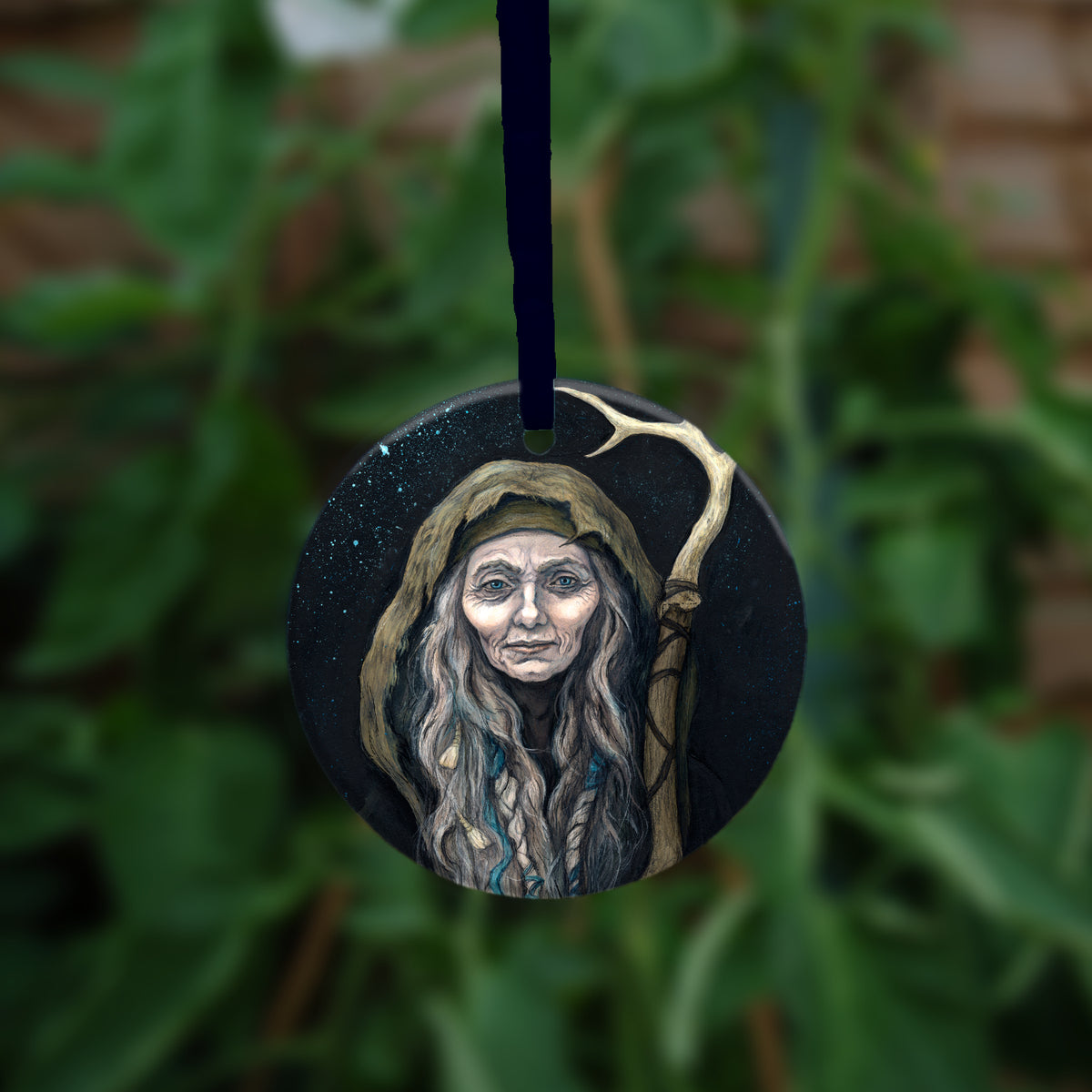 A circular item hanging against a leafy background: An older woman with blue eyes stands facing the viewer. Her head is covered by a brown hood, and her long grey hair, some of it braided with blue braid, falls down below her face. She holds a staff, topped with a deer antler, upright in one hand. A sea of stars shine behind her.