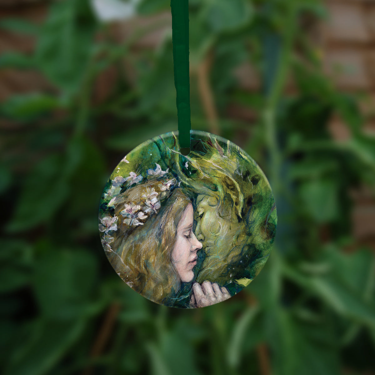 Circular hanging ornament with a green ribbon: A young woman with a garland of pale flowers in her hair closes her eyes as she moves to kiss a green man.