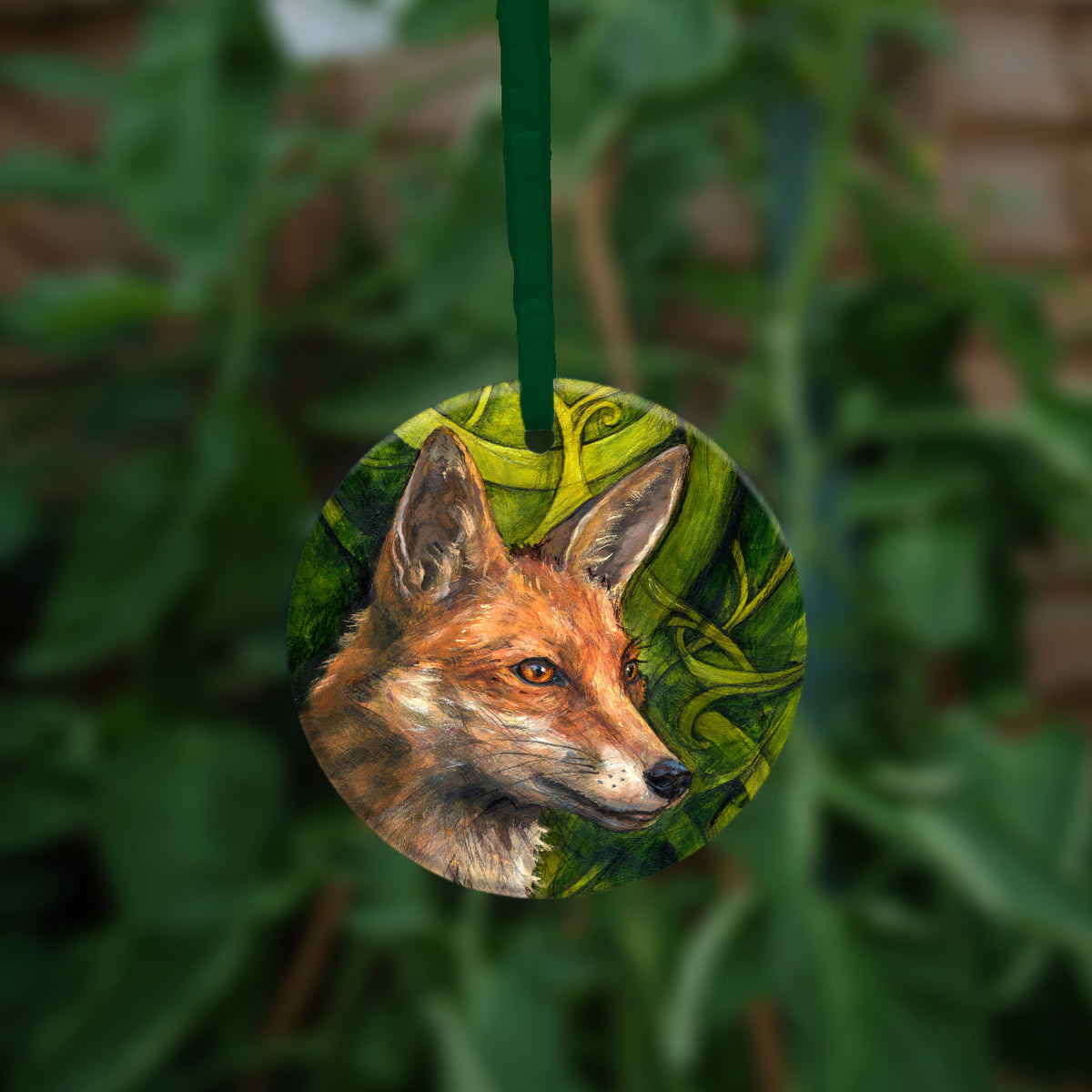 A circular hanging ornament: The head and neck of a red fox, their face angled towards the front right. The background consists of swirls of branches and leaves in a rich green colour.