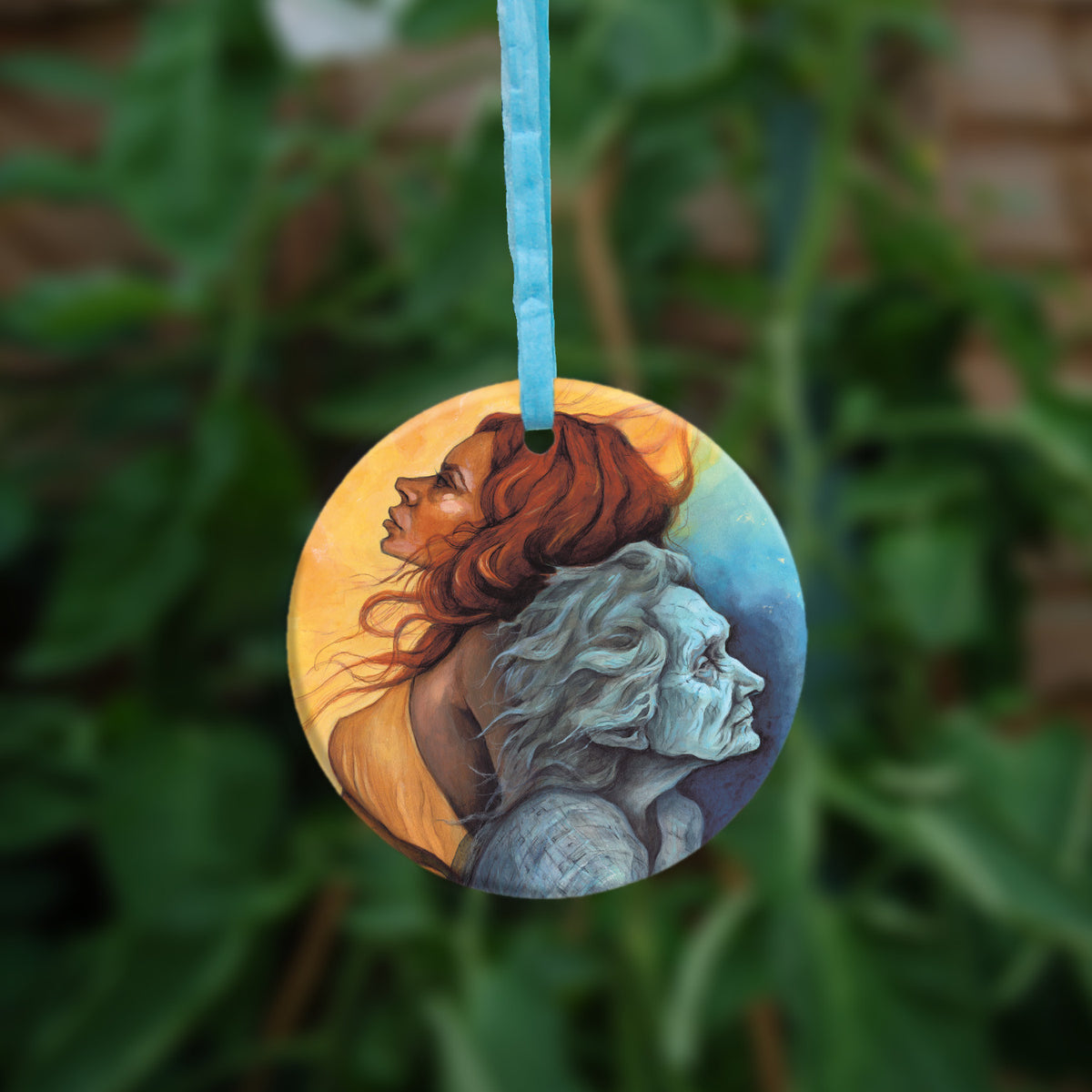 Circular hanging ornament with a light blue ribbon: A painting of two diagonal halves: to the top/left, against a background of mottled orange, a young woman with red hair and orange dress faces to the left. She appears to be leaning against, and blending with, an elderly woman who faces towards the right/bottom in the other half of the design. This half is coloured in shades of blue and grey.