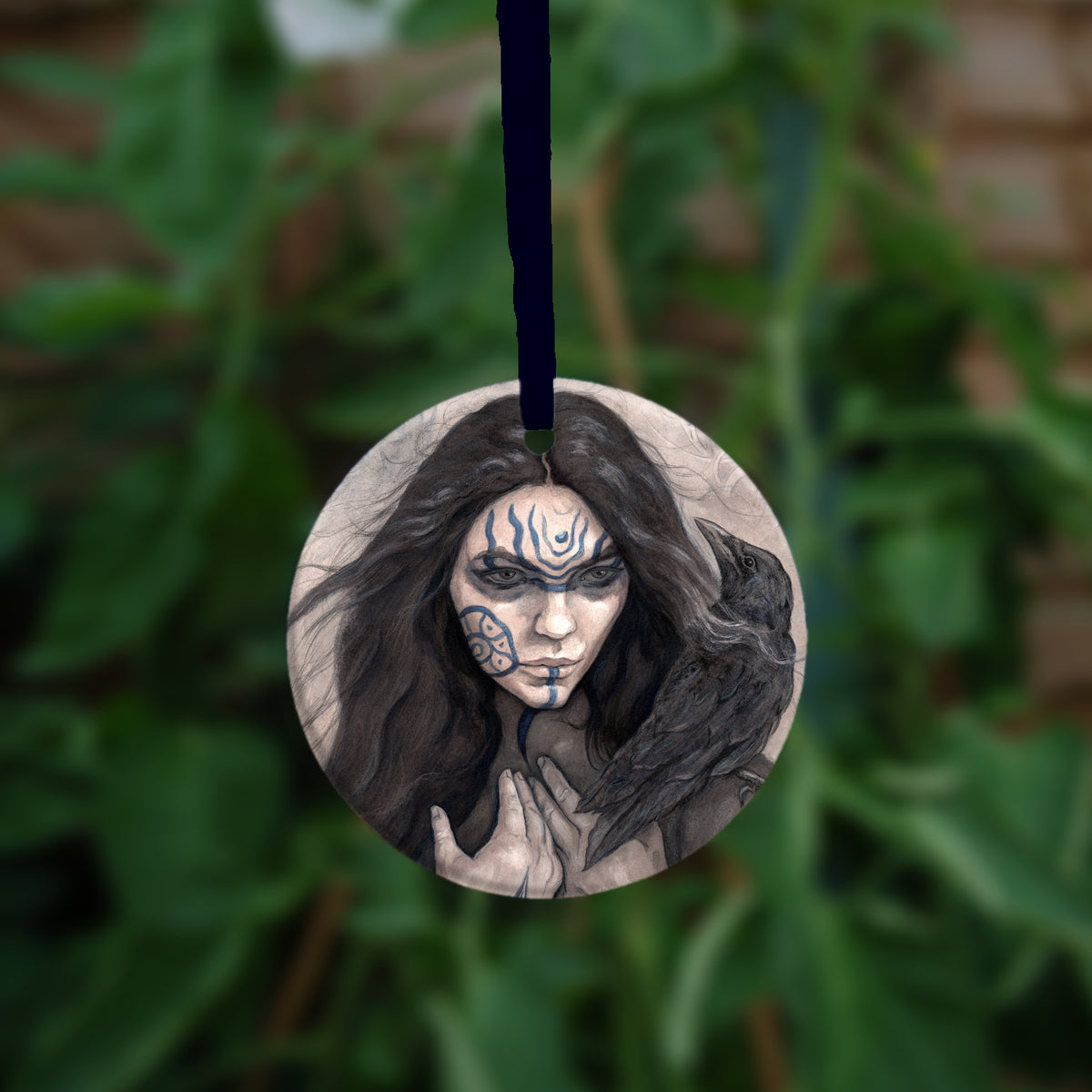 A circular hanging ornament: The head and shoulders of a woman with a raven sitting on her shoulder. She has long dark hair and her face is decorated with patterns in blue paint. Her hands are held together against her body. On her wrist she wears a blue band with a raven skull attached. The background is a mottled brown with feathers and swirls drawn within.