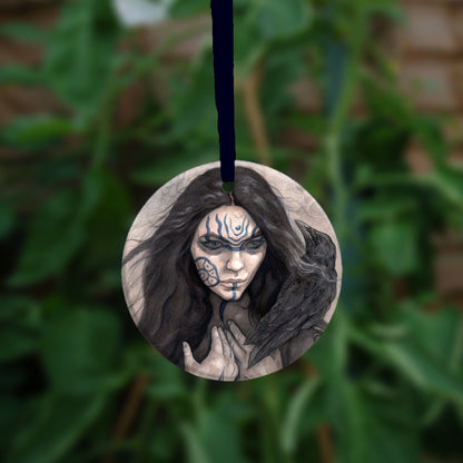 A circular hanging ornament: The head and shoulders of a woman with a raven sitting on her shoulder. She has long dark hair and her face is decorated with patterns in blue paint. Her hands are held together against her body. On her wrist she wears a blue band with a raven skull attached. The background is a mottled brown with feathers and swirls drawn within.