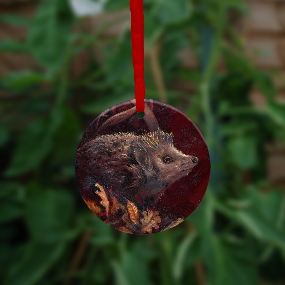 A hanging ornament on a red ribbon: A hedgehog shuffles through crisp brown autumn leaves lying on the ground. Woven branches rise upwards, and a leaf falls towards the ground. Colours are browns and reds.