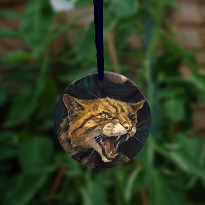A circular hanging ornament with a dark blue ribbon: The head and body of a snarling brown striped cat. 