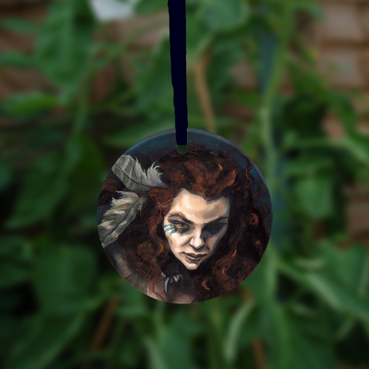 Circular hanging ornament with dark blue ribbon: The head and chest of a woman with long red hair decorated with two feathers, and with lines of blue paint extending away from her eyes.