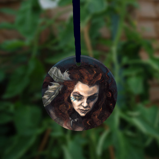 Circular hanging ornament with dark blue ribbon: The head and chest of a woman with long red hair decorated with two feathers, and with lines of blue paint extending away from her eyes.