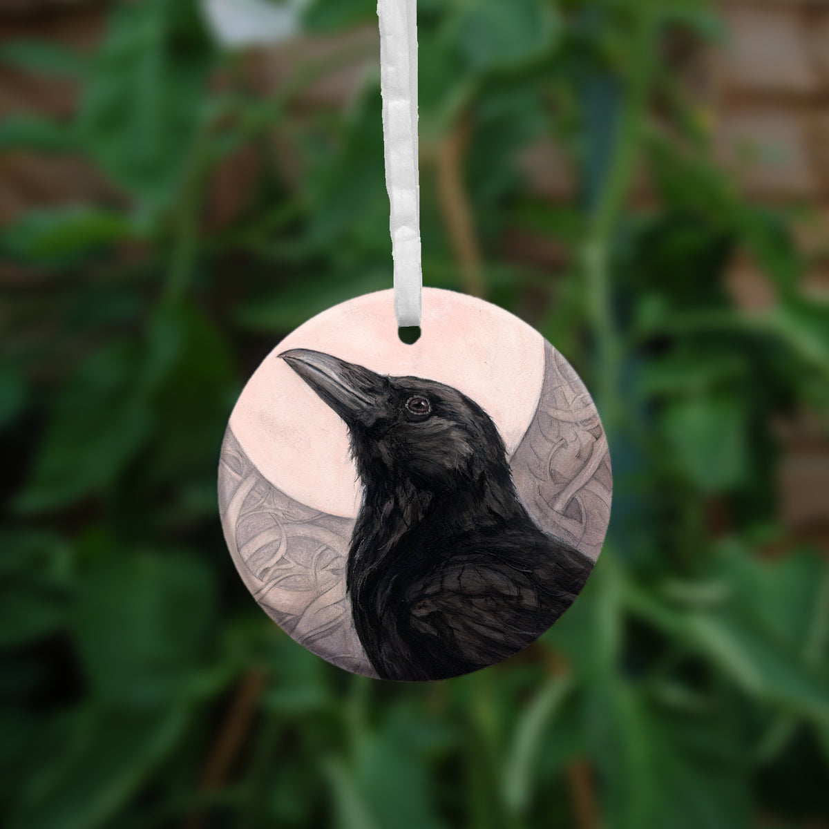 A circular hanging ornament with a white ribbon: The head and body of a raven standing facing towards the left, head tilted upward, behind which is a full moon. The background consists of swirls in a pale pink-brown colour.