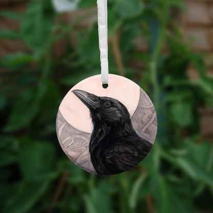 A circular hanging ornament with a white ribbon: The head and body of a raven standing facing towards the left, head tilted upward, behind which is a full moon. The background consists of swirls in a pale pink-brown colour.