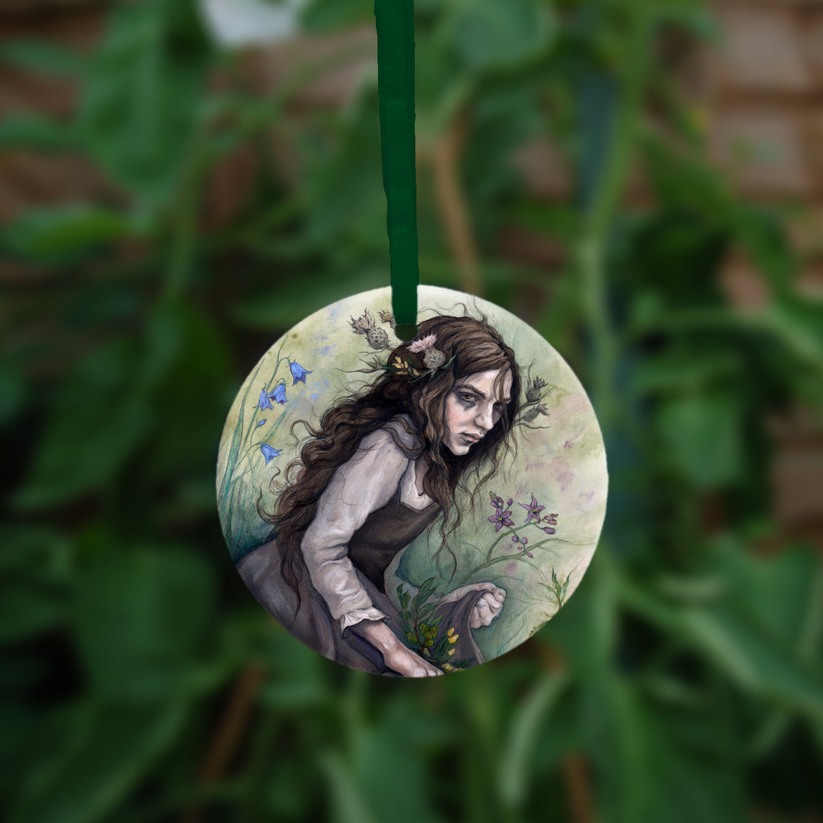 A circular hanging ornament with a green ribbon: A young woman wearing a long brown sleeveless dress with a pale long sleeved top underneath, is surrounded by flowers as she kneels on the ground. The front of her dress is gathered in her hands to form an apron containing sprigs of bog myrtle and yellow gorse flowers. Heather grows next to her. Behind her are harebells, with woody nightshade to the other side and nettles and yarrow to the front. Thistles appear to be tied into her long brown hair.
