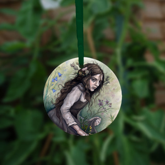 A circular hanging ornament with a green ribbon: A young woman wearing a long brown sleeveless dress with a pale long sleeved top underneath, is surrounded by flowers as she kneels on the ground. The front of her dress is gathered in her hands to form an apron containing sprigs of bog myrtle and yellow gorse flowers. Heather grows next to her. Behind her are harebells, with woody nightshade to the other side and nettles and yarrow to the front. Thistles appear to be tied into her long brown hair.