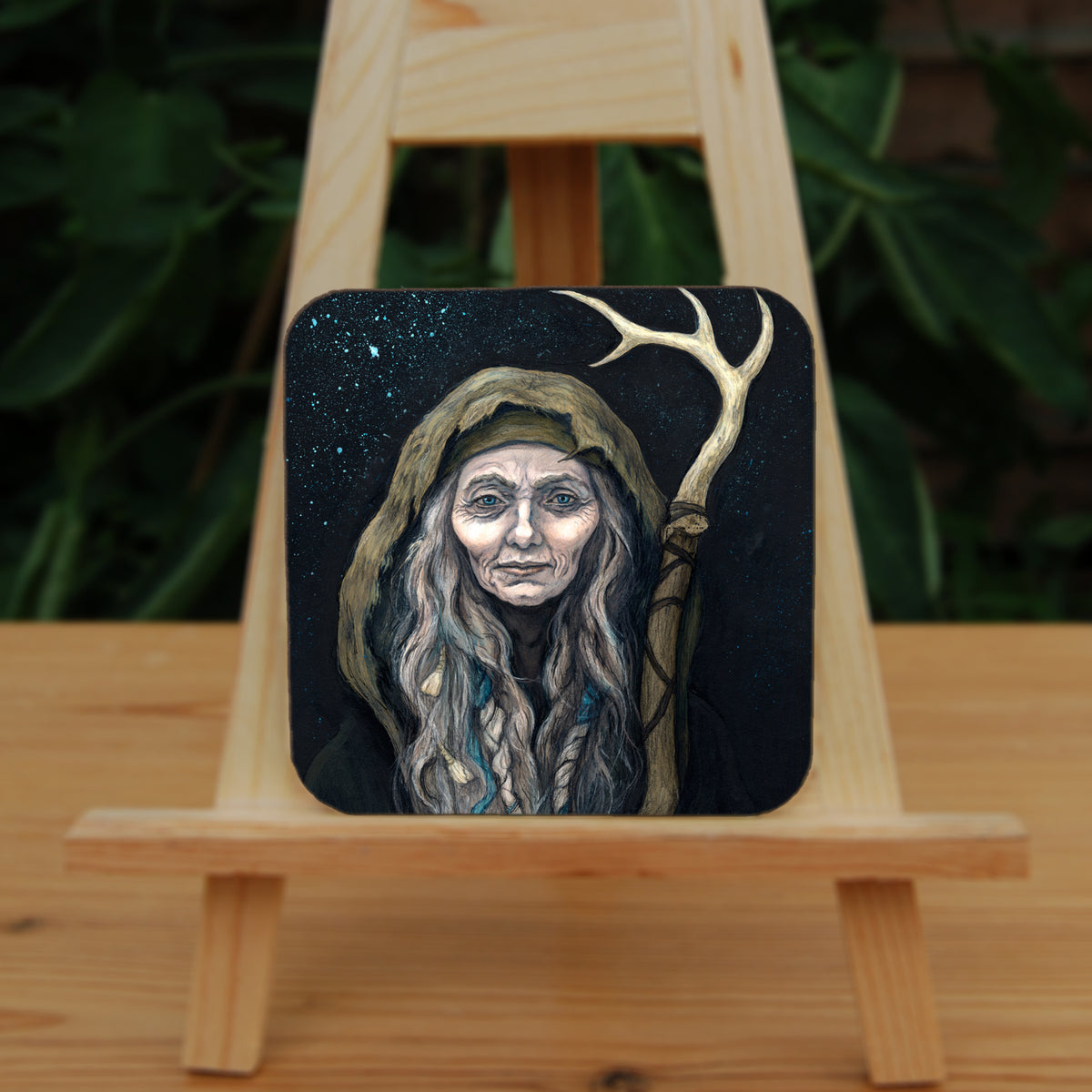 A square coaster: An older woman with blue eyes stands facing the viewer. Her head is covered by a brown hood, and her long grey hair, some of it braided with blue braid, falls down below her face. She holds a staff, topped with a deer antler, upright in one hand. A sea of stars shine behind her.
