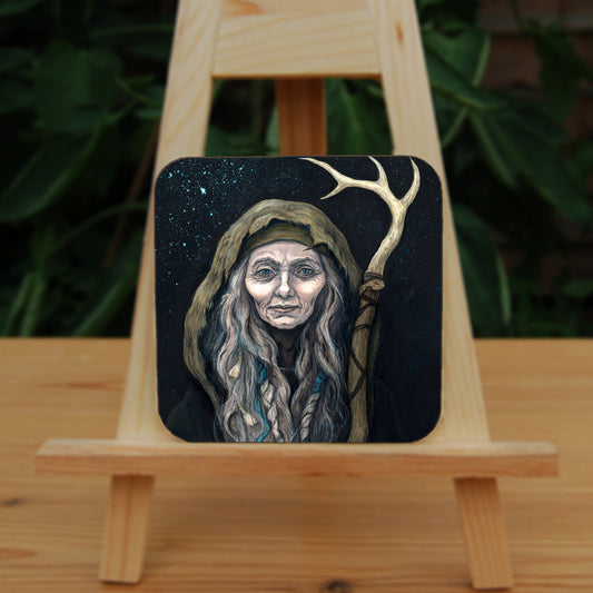 A square coaster: An older woman with blue eyes stands facing the viewer. Her head is covered by a brown hood, and her long grey hair, some of it braided with blue braid, falls down below her face. She holds a staff, topped with a deer antler, upright in one hand. A sea of stars shine behind her.