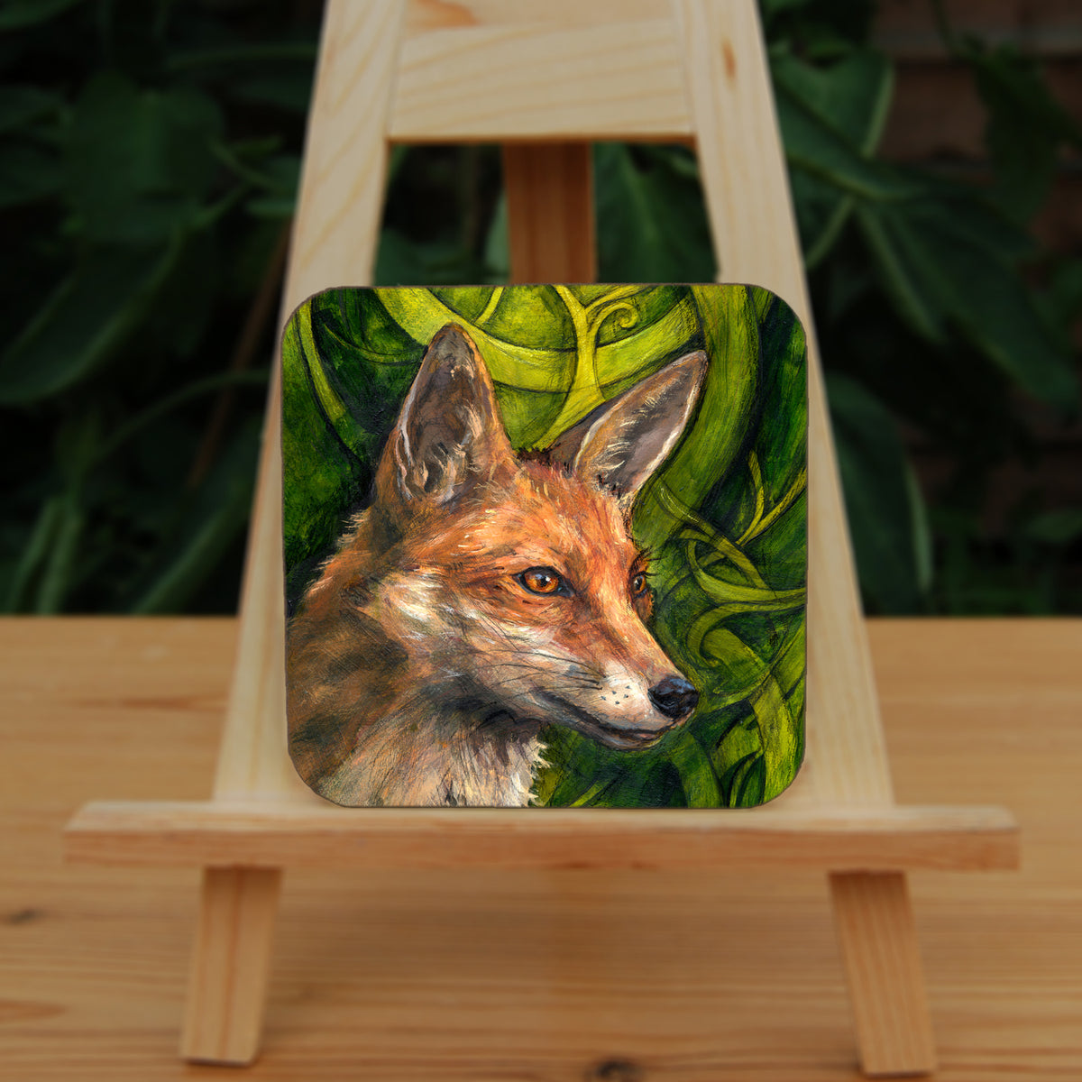 A square coaster: The head and neck of a red fox, their face angled towards the front right. The background consists of swirls of branches and leaves in a rich green colour.