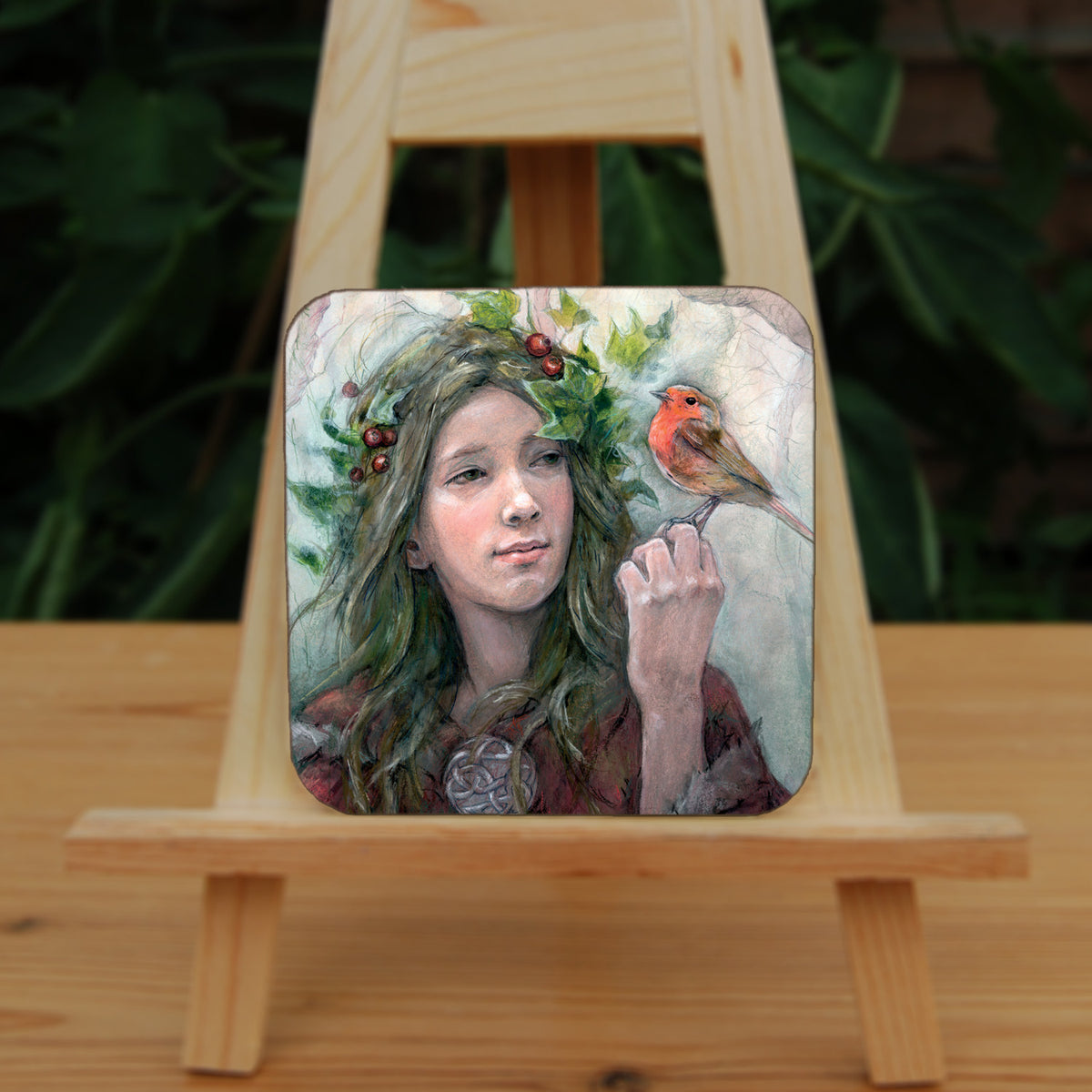 A square coaster: A young woman looks towards a robin sitting at eye level on the fingers of her hand. She wears a red cloak with white fur trim, closed with a large Celtic brooch. Sprigs of red berries and ivy are woven into her hair. 
