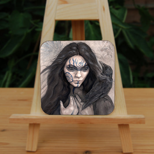 A square coaster: The head and shoulders of a woman with a raven sitting on her shoulder. She has long dark hair and her face is decorated with patterns in blue paint. Her hands are held together against her body. On her wrist she wears a blue band with a raven skull attached. The background is a mottled brown with feathers and swirls drawn within.