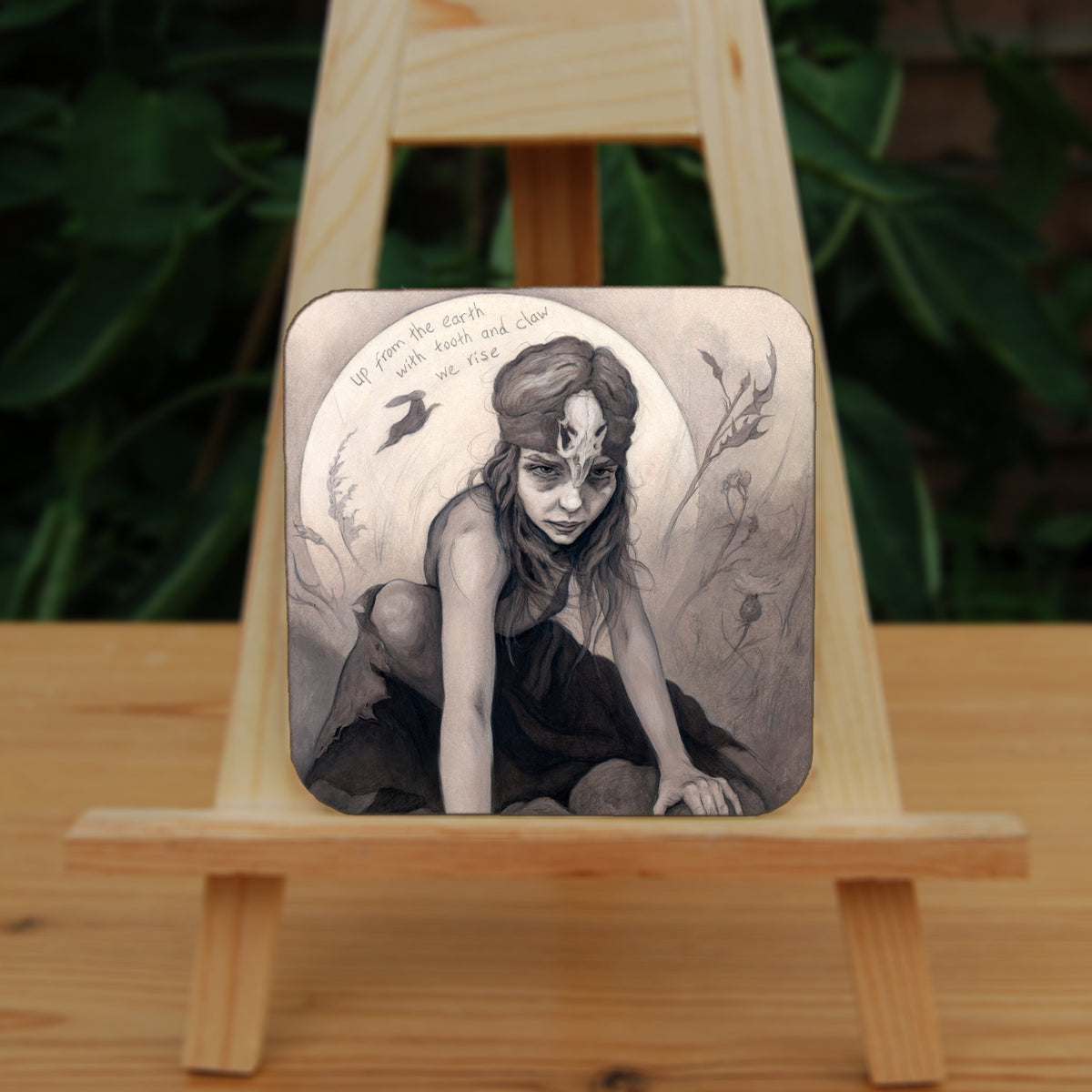 Square coaster: A young woman crouches on a pile of stones, her hands resting on stones in front of her. The woman wears a headband with a raven skull attached to the front. A full moon rises behind her, and a silhouette of a hare leaps in front of it. An inscription over the moon reads: Up from the earth with tooth and claw we rise. Stems and flowers fill the rest of the scene.