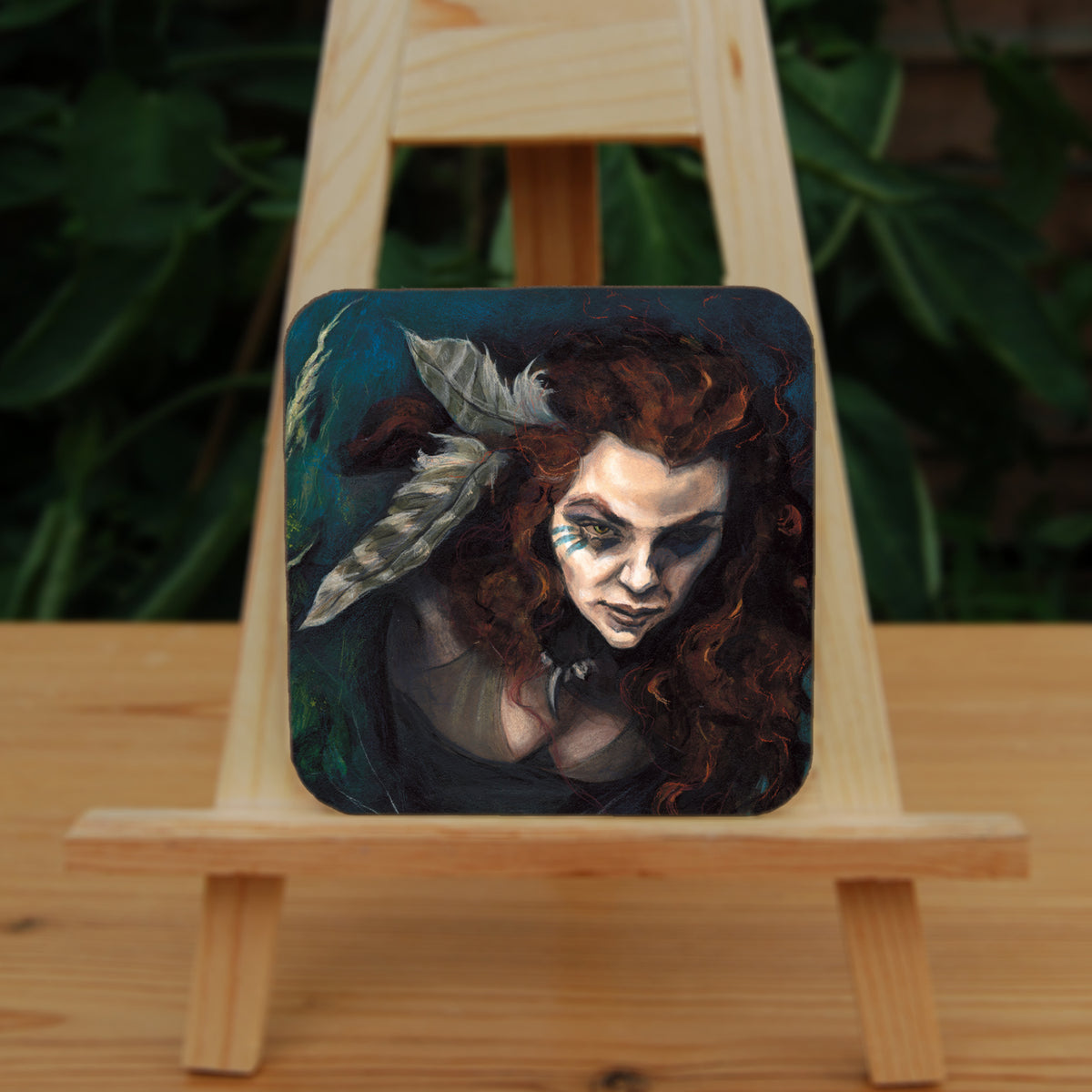 A square coaster: The head and chest of a woman with long red hair decorated with two feathers, and with lines of blue paint extending away from her eyes. The background is teal and green vegetation.