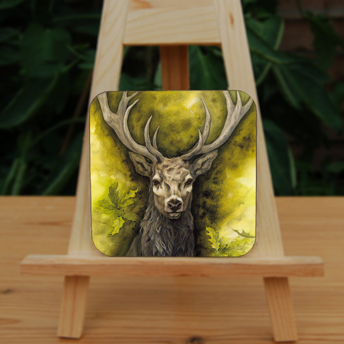 A square coaster: The head and chest of an antlered stag directly facing the viewer. Oak leaves are drawn to his left and right. The background is mottled yellows and browns.