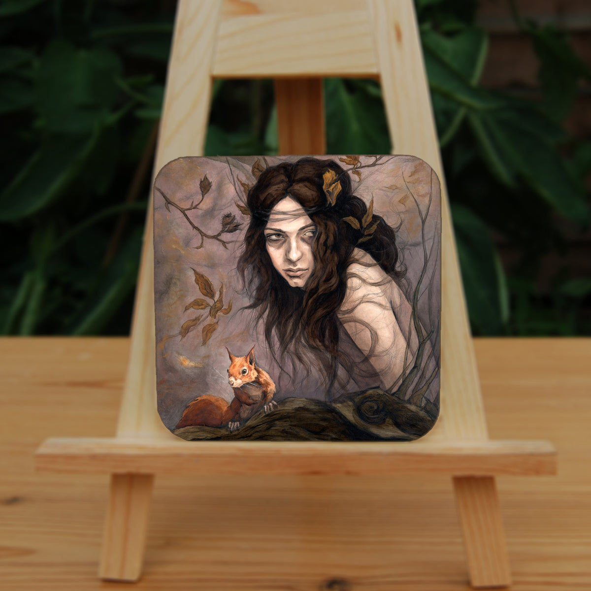 A square coaster: The head and body of a young woman appears above a thick tree branch, upon which sits a red squirrel. The woman is turning her head as if to look behind her. She has long brown hair with leaves stuck in it. Autumnal leaves and branches fill the scene. Colours are reds, oranges and browns.