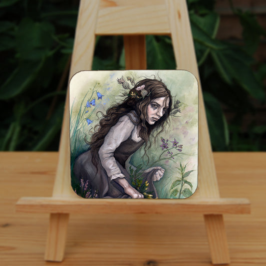 A square coaster: A young woman wearing a long brown sleeveless dress with a pale long sleeved top underneath, is surrounded by flowers as she kneels on the ground. The front of her dress is gathered in her hands to form an apron containing sprigs of bog myrtle and yellow gorse flowers. Heather grows next to her. Behind her are harebells, with woody nightshade to the other side and nettles and yarrow to the front. Thistles appear to be tied into her long brown hair.