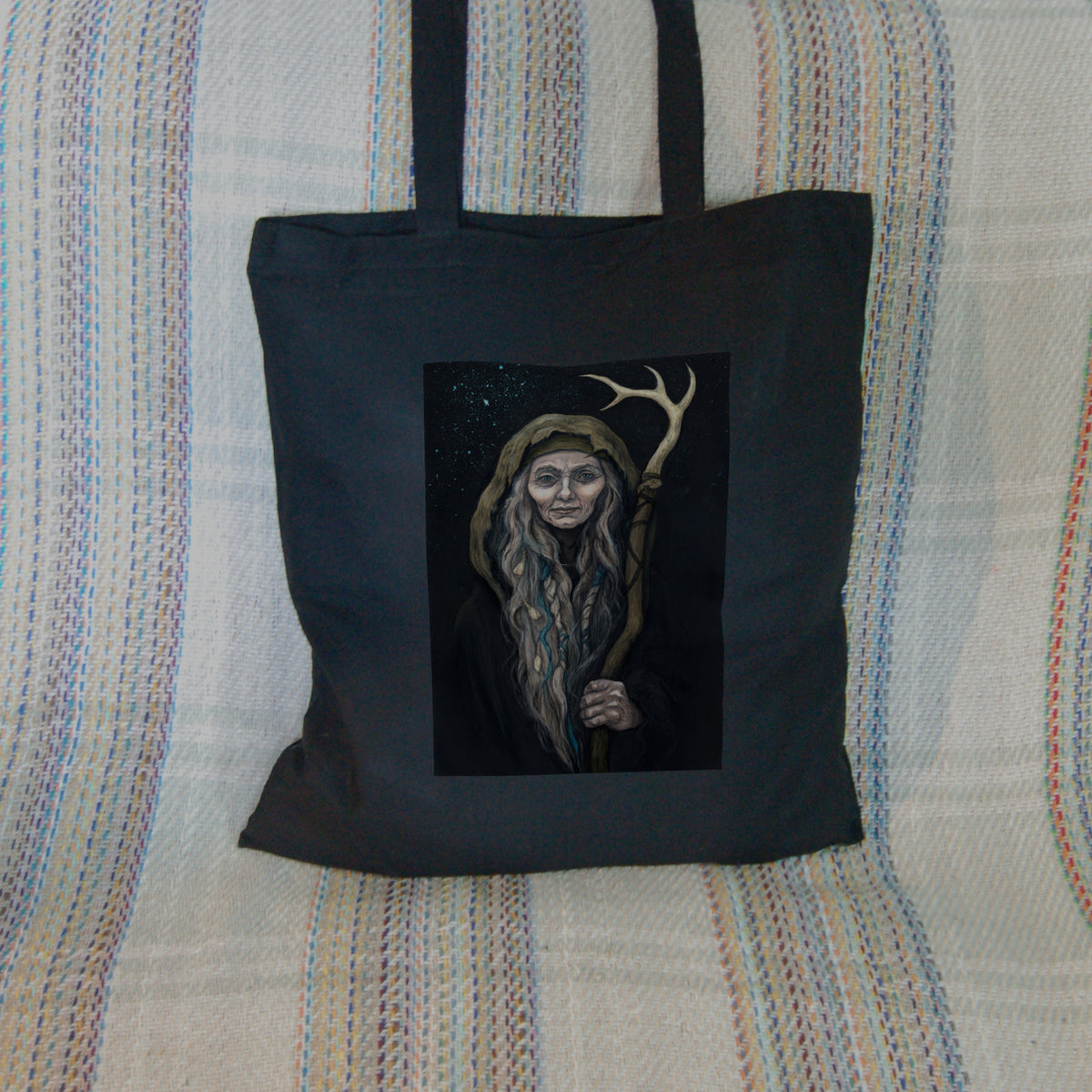 Black tote bag: An older woman with blue eyes stands facing the viewer. Her head is covered by a brown hood, and her long grey hair, some of it braided with blue braid, falls down below her face. She holds a staff, topped with a deer antler, upright in one hand. A sea of stars shine behind her.