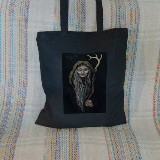 Black tote bag: An older woman with blue eyes stands facing the viewer. Her head is covered by a brown hood, and her long grey hair, some of it braided with blue braid, falls down below her face. She holds a staff, topped with a deer antler, upright in one hand. A sea of stars shine behind her.