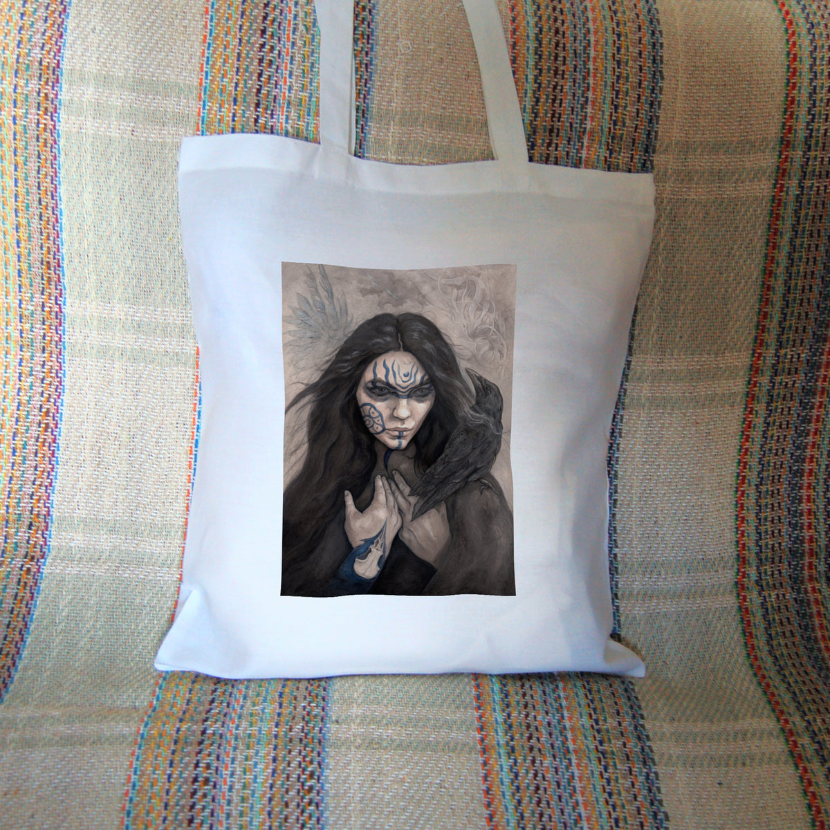 White tote bag: The head and shoulders of a woman with a raven sitting on her shoulder. She has long dark hair and her face is decorated with patterns in blue paint. Her hands are held together against her body. On her wrist she wears a blue band with a raven skull attached. The background is a mottled brown with feathers and swirls drawn within.