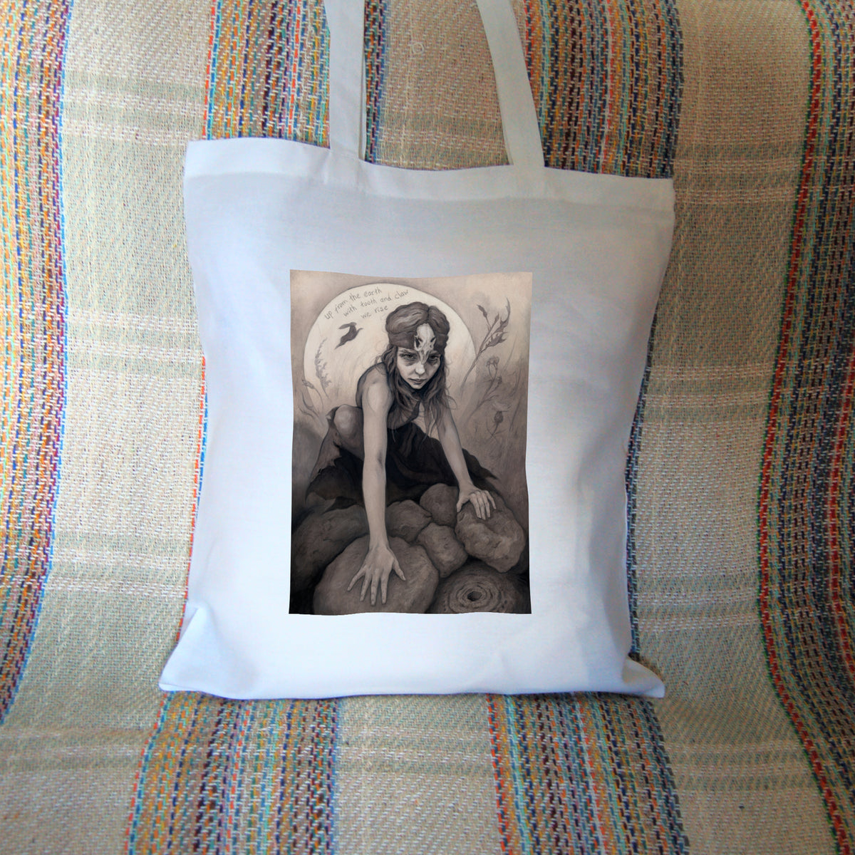 White tote bag: A young woman crouches on a pile of stones, her hands resting on stones in front of her. In front of the stones, a little lower down, is a stone with a round hole in it surrounded by a circular pattern. The woman wears a headband with a raven skull attached to the front. A full moon rises behind her, and a silhouette of a hare leaps in front of it. An inscription over the moon reads: Up from the earth with tooth and claw we rise. Stems and flowers fill the rest of the scene.