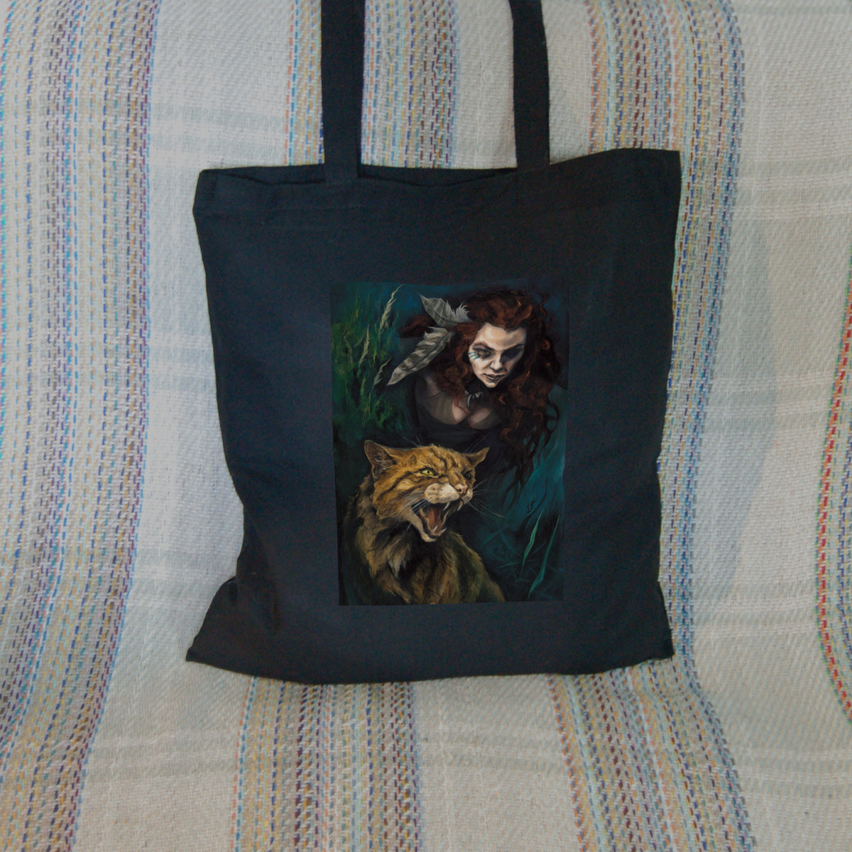 Black tote bag: The head and chest of a woman with long red hair decorated with two feathers, and with lines of blue paint extending away from her eyes, looms into the picture above the head and body of a snarling brown striped cat. The background is teal and green vegetation.