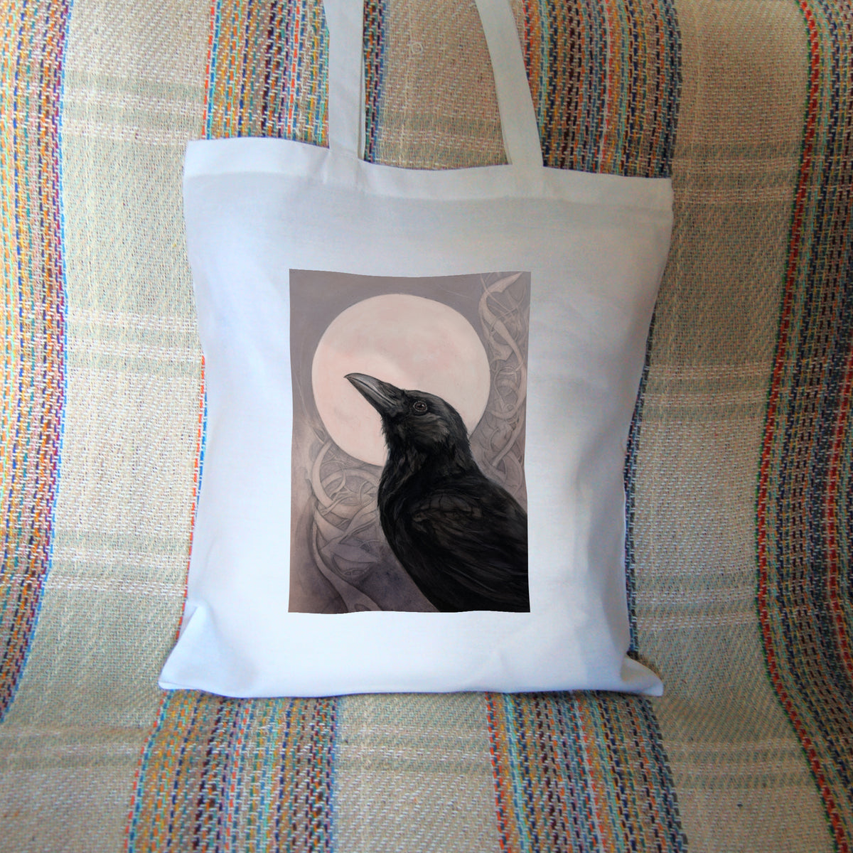 White tote bag: The head and body of a raven standing facing towards the left, head tilted upward, behind which is a full moon. The background consists of swirls in a pale pink-brown colour.