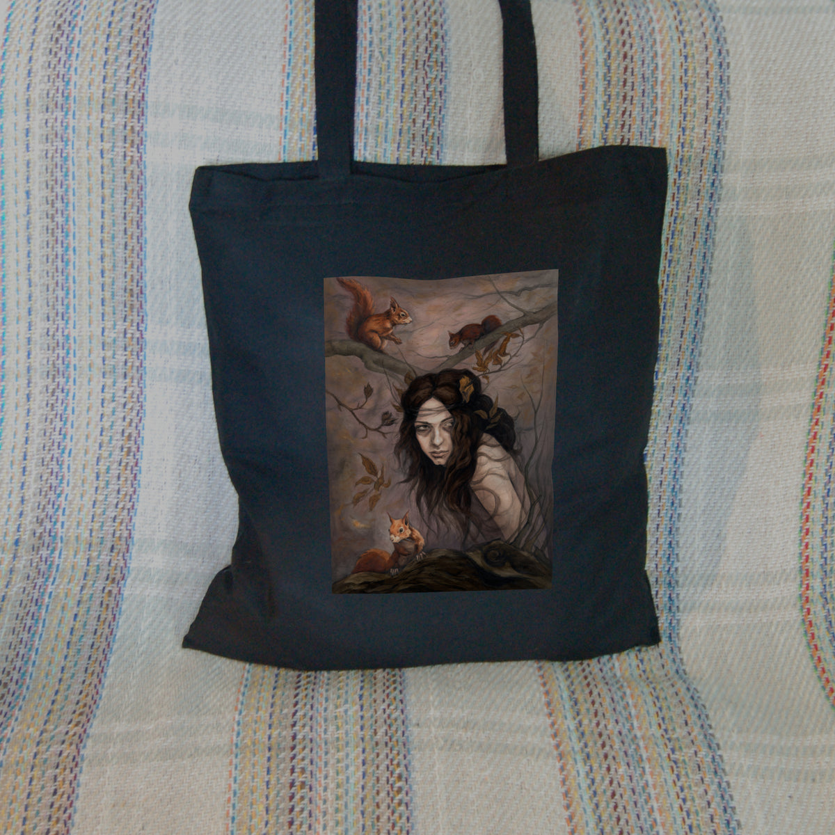 Black tote bag: The head and body of a young woman appears above a thick tree branch, upon which sits a red squirrel. Two more squirrels crouch on a branch above her. The woman is turning her head as if to look behind her. She has long brown hair with leaves stuck in it. Autumnal leaves and branches fill the scene. Colours are reds, oranges and browns.