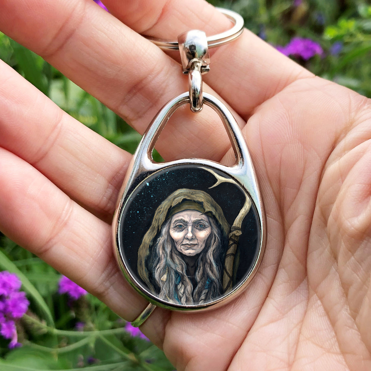 A hand holding an oval metal keyring with a circular central image: An older woman with blue eyes stands facing the viewer. Her head is covered by a brown hood, and her long grey hair, some of it braided with blue braid, falls down below her face. She holds a staff, topped with a deer antler, upright in one hand. A sea of stars shine behind her.