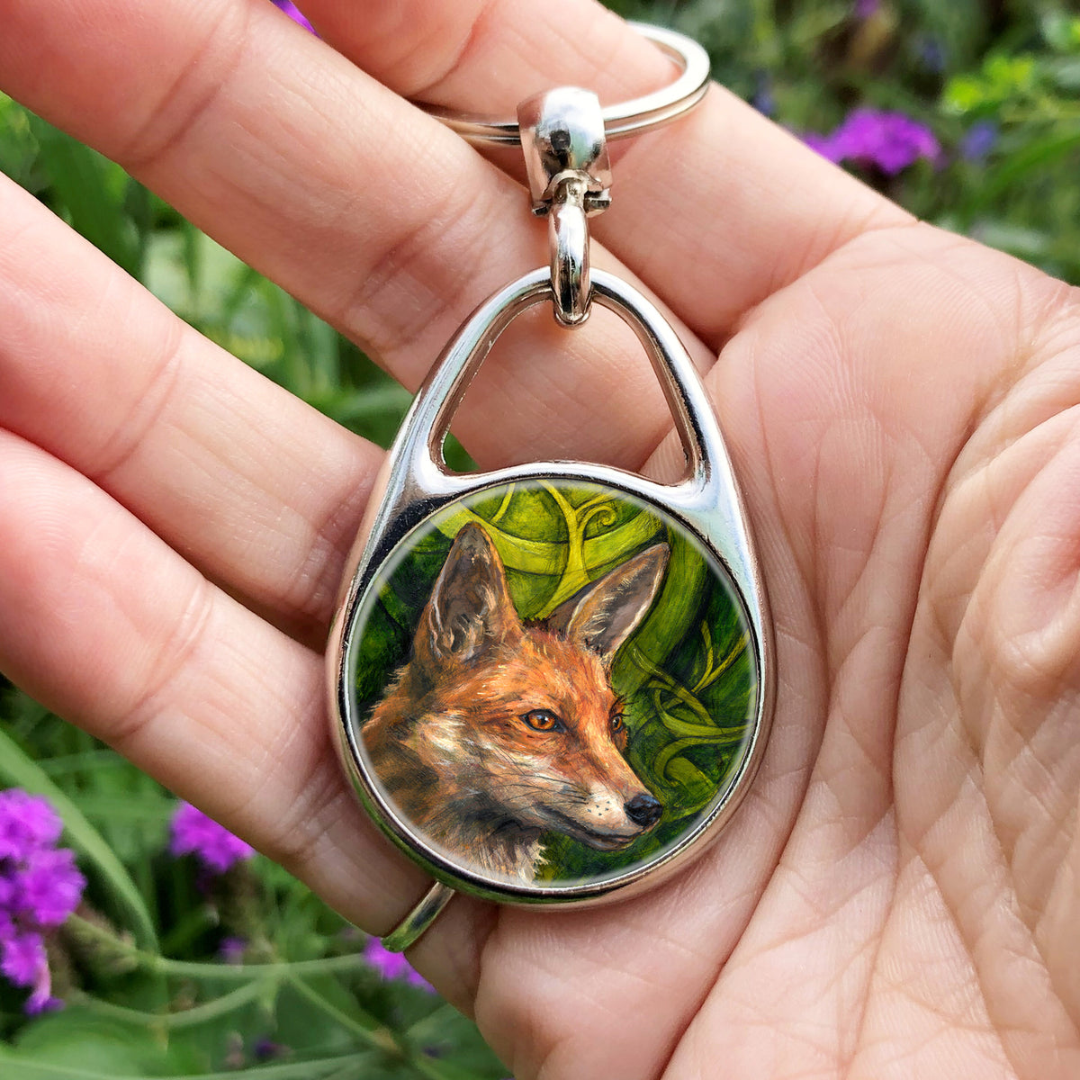 A hand holding an oval keyring with a central circular image: The head and neck of a red fox, their face angled towards the front right. The background consists of swirls of branches and leaves in a rich green colour.