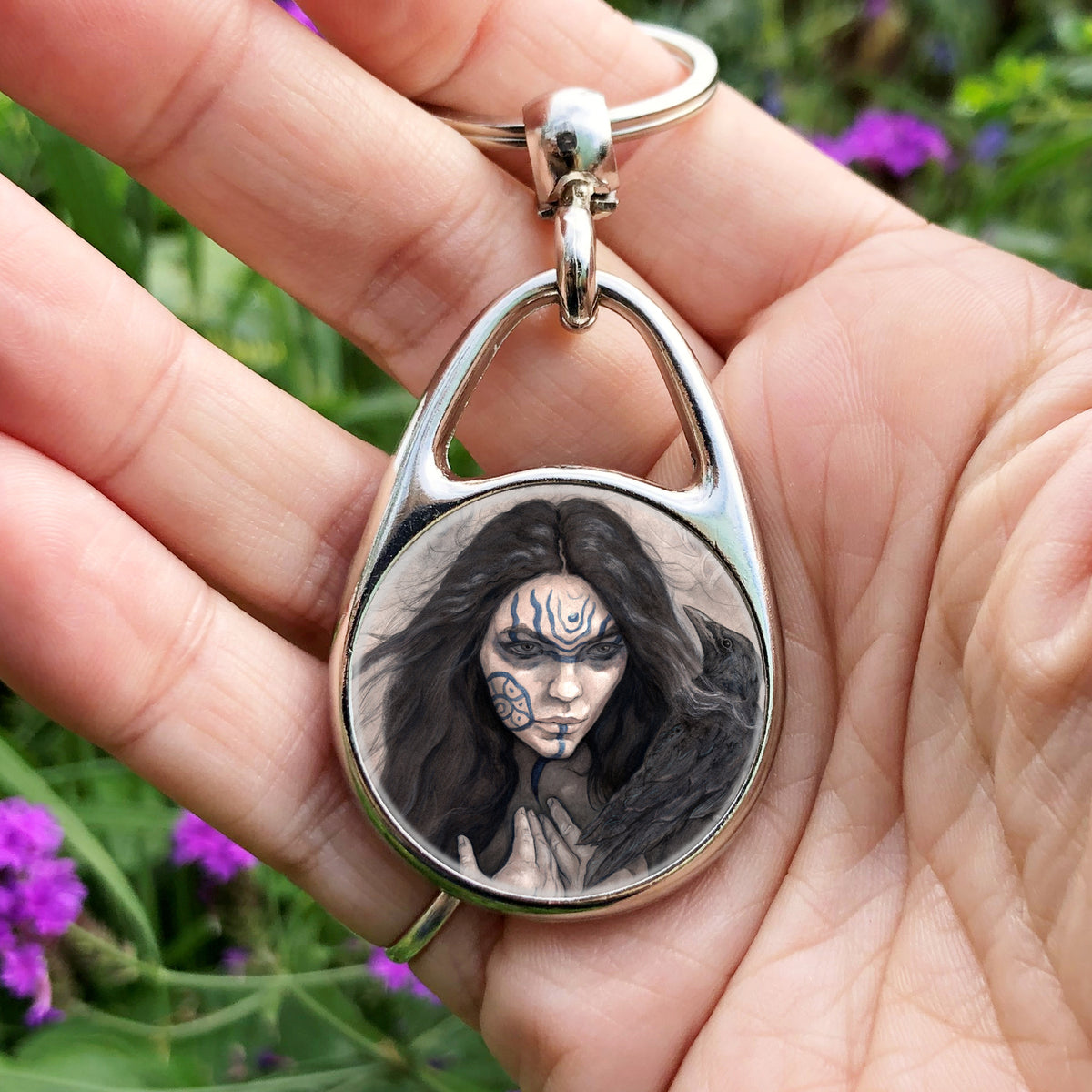 A hand holding an oval keyring with a circular central image: The head and shoulders of a woman with a raven sitting on her shoulder. She has long dark hair and her face is decorated with patterns in blue paint. Her hands are held together against her body. On her wrist she wears a blue band with a raven skull attached. The background is a mottled brown with feathers and swirls drawn within.
