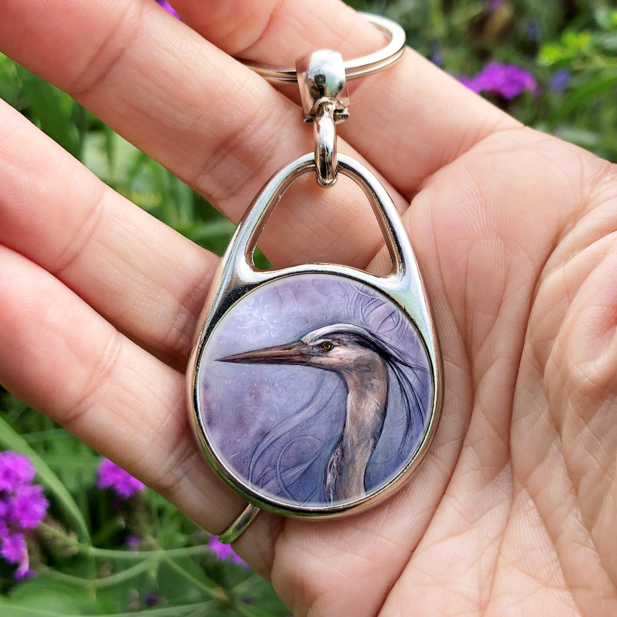 A hand holding an oval metal keyring with a circular inset design: A sideways view of the head and neck of a heron. The background is a mottled purple with a swirl pattern.