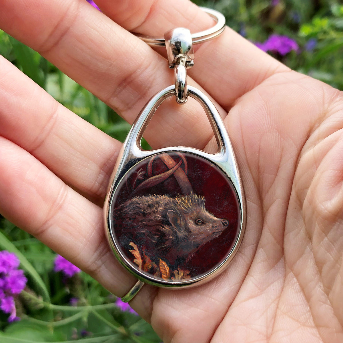 A hand holding an oval metal keyring with a circular insert: A hedgehog shuffles through crisp brown autumn leaves lying on the ground. Woven branches rise upwards, and a leaf falls towards the ground. Colours are browns and reds.