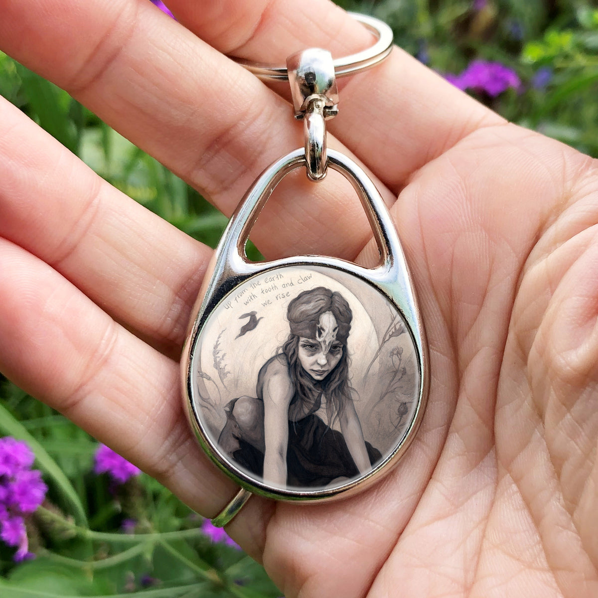 A hand holding an oval metal keyring with a circular insert: A young woman crouches on a pile of stones, her hands resting on stones in front of her. The woman wears a headband with a raven skull attached to the front. A full moon rises behind her, and a silhouette of a hare leaps in front of it. An inscription over the moon reads: Up from the earth with tooth and claw we rise.