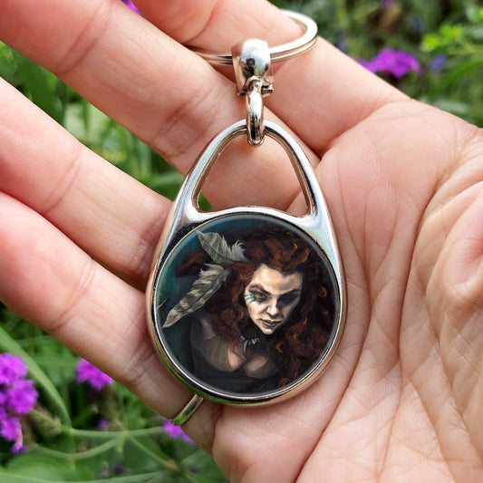 A hand holding an oval metal keyring with a circular insert: The head and chest of a woman with long red hair decorated with two feathers, and with lines of blue paint extending away from her eyes.