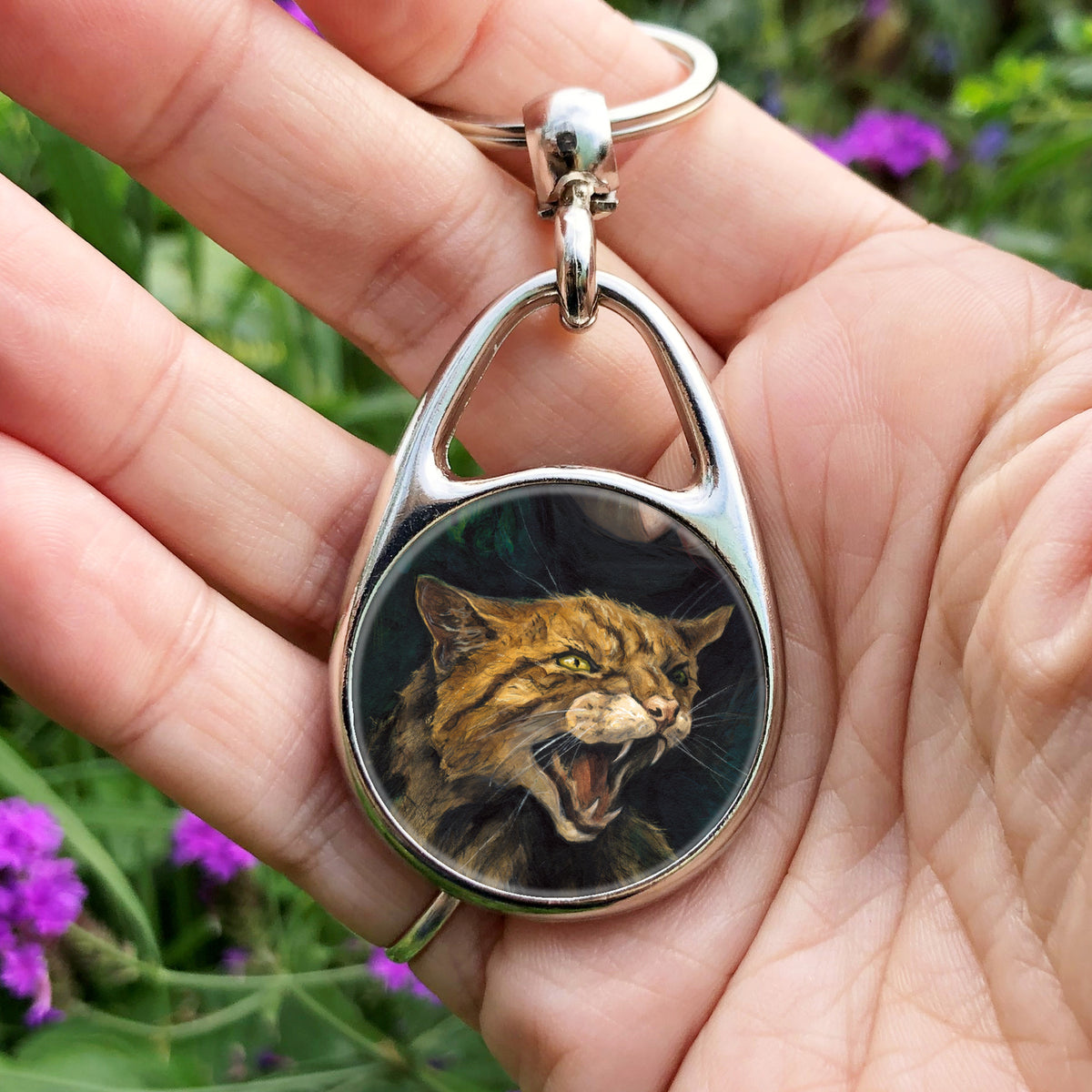 A hand holding an oval metal keyring with a circular insert: The head and body of a snarling brown striped cat. 